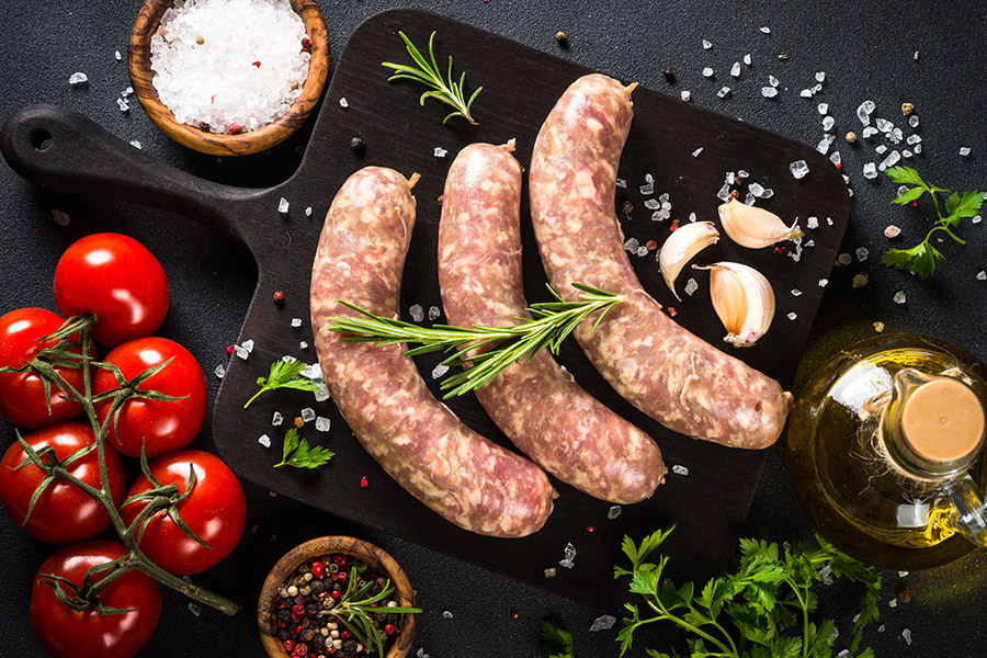 Regionale Bratwurst Drei rohe Würste auf Schneidebrett mit Tomaten, Knoblauch, Kräutern und grobem Salz.